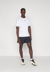 Λευκό t-shirt με στρογγυλή λαιμόκοψη και μαύρο λογότυπο στο στήθος, σε συνδυασμό με μαύρο σορτς. Το μοντέλο φοράει λευκές κάλτσες και αθλητικά παπούτσια σε μαύρο και κρεμ χρώμα.