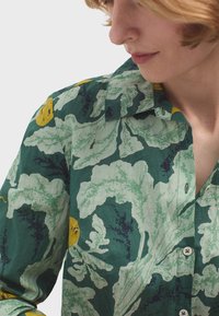 Camicia verde floreale con grandi motivi astratti di foglie bianche e gialle, realizzata in tessuto leggero con colletto classico e chiusura a bottoni.
