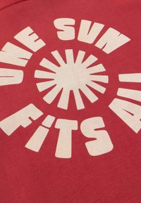 Tissu rouge avec un texte beige lisant "ONE SUN FITS ALL" disposé en cercle autour d'une forme de soleil stylisée au centre.
