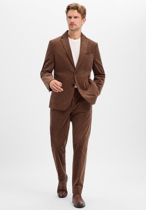 Man walking wearing a brown corduroy suit with a white shirt and brown leather shoes on a plain light background.