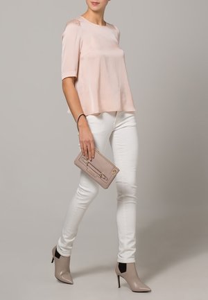 Blouse en satin rose clair à manches courtes, associée à un jean blanc slim. Le modèle tient une pochette rose clair et porte des escarpins beiges à bouts pointus.