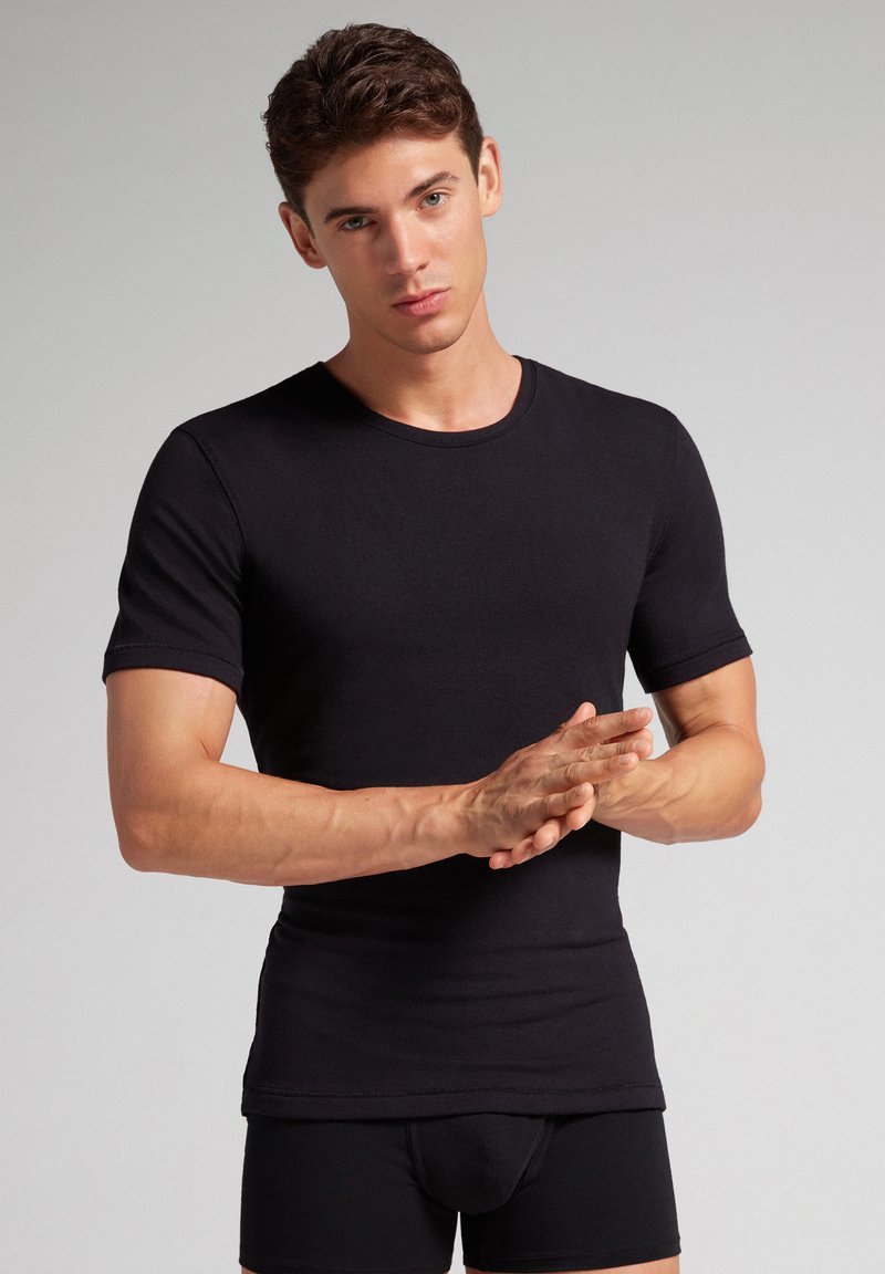 Camiseta de algodón negra de manga corta con cuello redondo, forma ajustada, textura suave y detalles de costura mínimos. El modelo posa con las manos juntas.