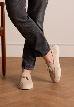 Beige suede slip-on shoes with a rounded toe and decorative bow detail, paired with dark denim jeans, on a wooden floor.