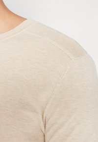 Suéter de malha bege claro com uma textura suave e gola canelada. Apresenta um detalhe de malha subtil na costura do ombro.