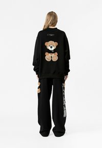 Sudadera negra de gran tamaño con un bordado de un oso de peluche en la parte posterior, combinada con pantalones negros, que también cuentan con un detalle de oso de peluche.