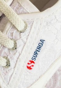 Sneaker bianca in tela con tessuto testurizzato, occhielli metallici rotondi e lacci bianchi. Presenta un logo colorato "SUPERGA" sul lato.