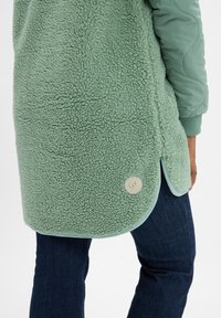 Gros plan sur une personne portant une veste en polaire sherpa verte avec des manches matelassées et un jean bleu foncé, la main reposant le long du corps.