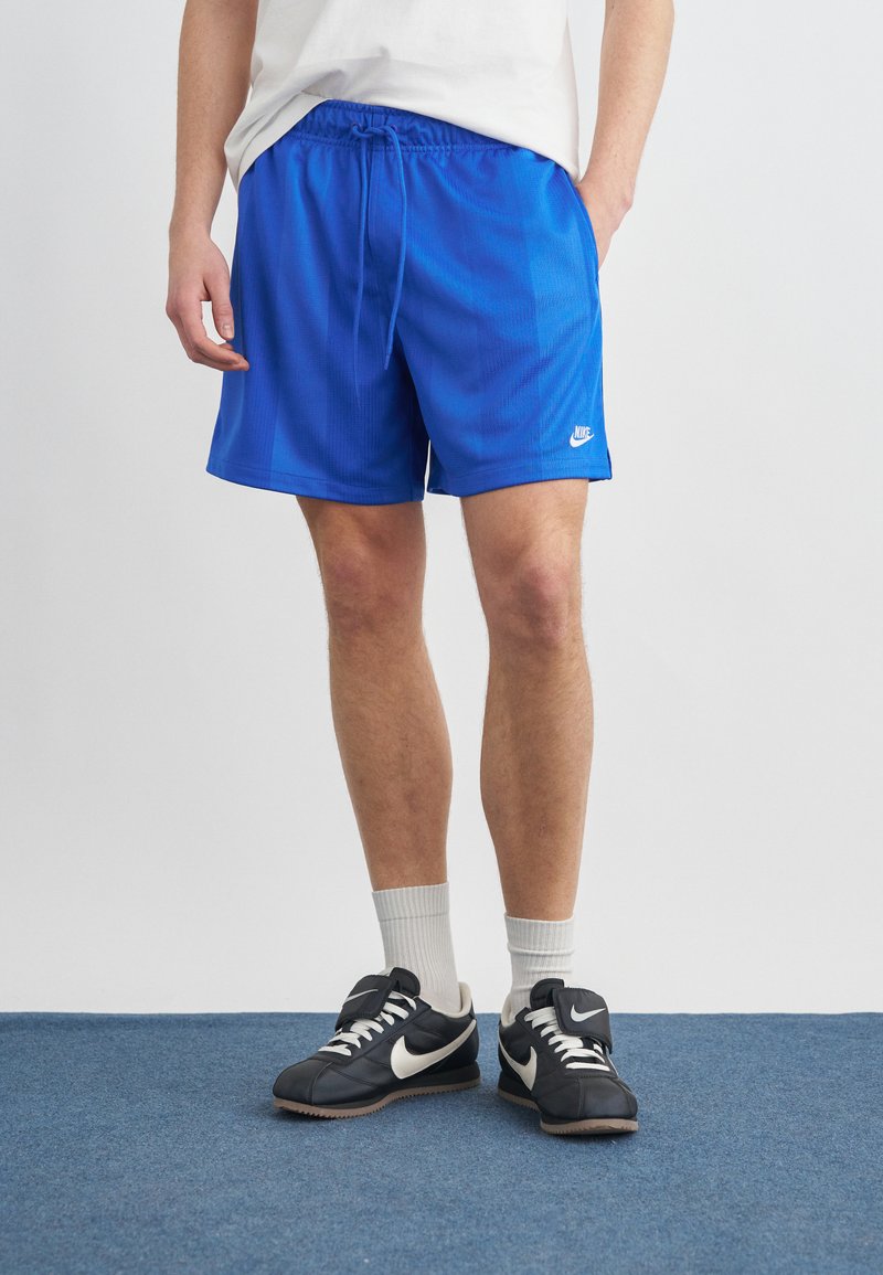 Άνδρας που φορά μπλε σορτς Nike, λευκές κάλτσες, μαύρα αθλητικά παπούτσια Nike, στέκεται σε μπλε χαλί με το ένα χέρι στην τσέπη.