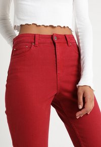 Jean en denim rouge à taille haute, doté d'une fermeture classique par bouton et fermeture éclair, d'un design à cinq poches, et d'une finition en tissu texturé.