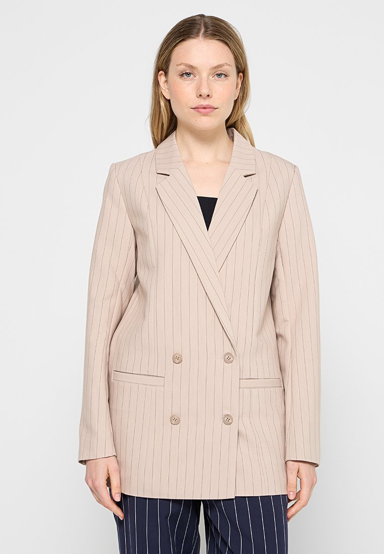 Femme portant un blazer beige à double boutonnage avec de fines rayures verticales et un pantalon à rayures marines, se tenant devant un fond clair.
