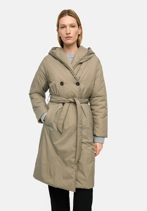 Kobieta nosząca beżowy, pikowany płaszcz sięgający do kolan, z paskiem, kapturem i podwójnymi czarnymi guzikami, stojąca z jedną ręką w kieszeni.