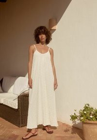 Robe maxi blanche texturée avec des bretelles spaghetti, présentant une coupe ample et un design superposé. Associée à des tongs rouges.