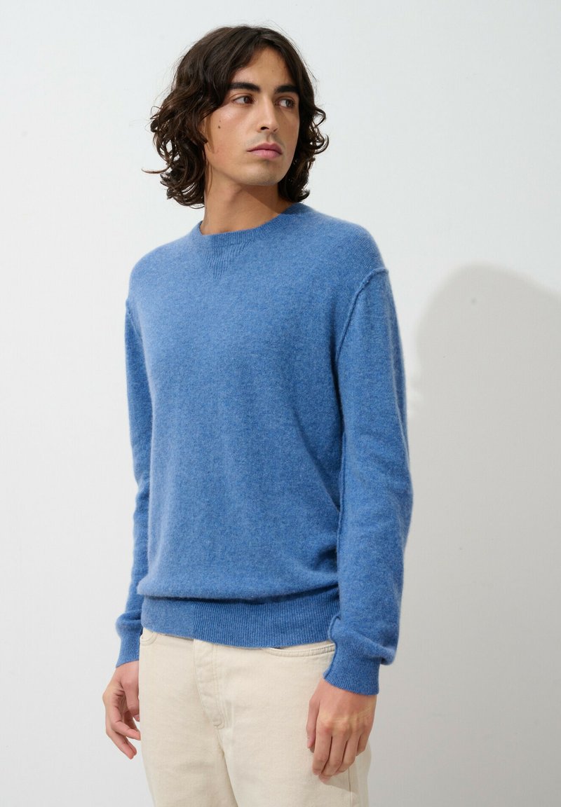 Hellblauer Wollpullover mit Rundhalsausschnitt, langen Ärmeln und geripptem Saum. Weicher Stoff und lockere Passform, kombiniert mit beigen Hosen.