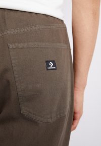 Smeđe cargo hlače sa stražnjim džepom s oznakom crnog Converse loga. Tkanina ima mekan, teksturiran završetak i vidljive detalje šivanja.