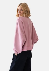 Różowa, oversize'owa bluza z okrągłym dekoltem, obniżonymi ramionami i ściągaczem na dole. Noszona z ciemnymi spodniami, prezentując luźny krój i miękką fakturę.