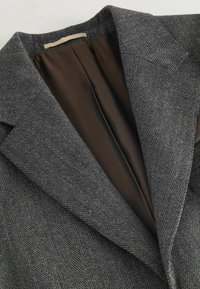 Grauer Fischgrat-Blazer mit einem spitz zulaufenden Revers, einem braunen Innenfutter und strukturierten Schultern. Strukturierter Stoff mit maßgeschneiderter Passform.