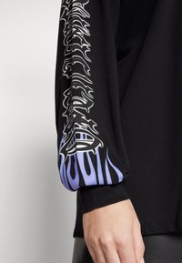 Svart långärmad tröja med grafiskt tryck som har vita mönster och lila låga accenter på ärmarna. Mjuk, bomullsmaterial.