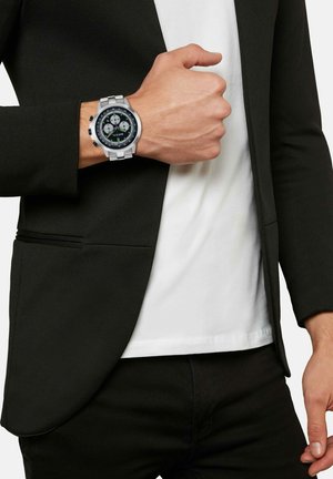 Montre multi-fonction argentée avec un cadran noir et vert, des accents en caoutchouc et un bracelet en acier inoxydable, associée à un blazer noir et une chemise blanche.