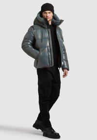 Szara kurtka puchowa z połyskującym wykończeniem, oversize'owym kapturem, zamkiem z przodu i elastycznymi mankietami, zestawiona z czarnymi spodniami typu cargo i butami sznurowanymi.