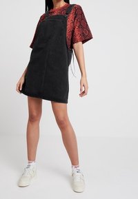 Svart denimoverallklänning med justerbara remmar, en framficka och rak skärning. Kombinerad med en röd t-shirt med reptilmönster och vita sneakers.