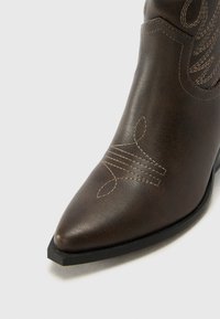 Hnědá kožená kovbojská bota s špičatou špičkou, vyšívanými detaily a matným povrchem. Pevná černá podrážka s mírným podpatkem.