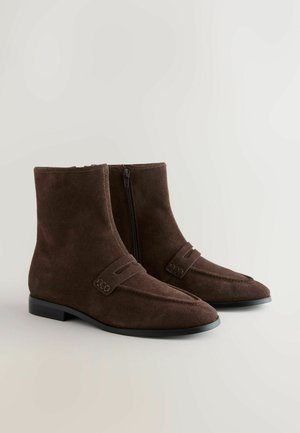 Botine din suede maro, cu vârf ascuțit, închidere cu fermoar și detalii decorative cu curea. Au o talpă plată de culoare neagră.