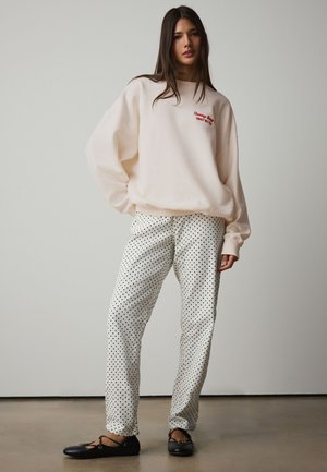 Noor naine kannab koorevärvi ülepõlve dressipluusi punase kirjaga, valgeid täpilisi pükse ja musti baleriine ning seisab ühevärvilise tausta ees.