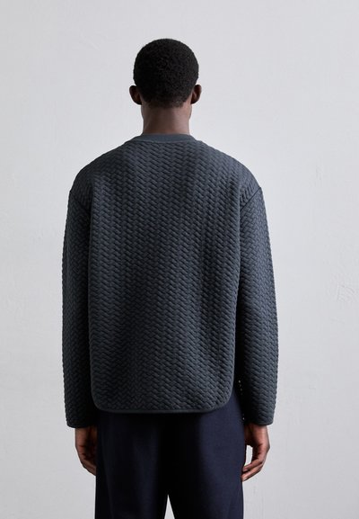 Pull en gris foncé, texturé, avec un motif tressé, un col rond et une coupe ample, associé à un pantalon foncé, vu de dos.