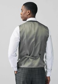 Gilet en satin gris avec une texture lisse, doté d'une sangle ajustable au dos, associé à une chemise blanche et un pantalon à carreaux.