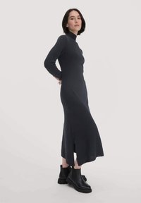 Langärmliges, dunkelgraues Kleid mit einem Stehkragen, aus Strickstoff gefertigt, mit figurbetontem Schnitt und ausgestelltem Saum. Kombiniert mit schwarzen Knöchelstiefeln.