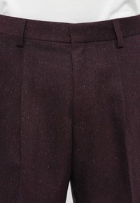 Dark burgundy speckled wool trousers with belt loops and front pleats, worn with a white top.