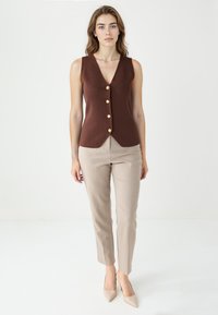 Kobieta stoi, ubrana w bezrękawnik w brązowym kolorze z guzikami, beżowe spodnie i beżowe szpilki na tle jednolitego białego tła.