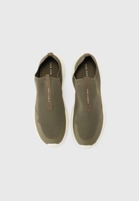 Sneakers slip-on verde oliva con tomaia in rete, suola in gomma crema e un tassello in pelle beige con testo sulla parte superiore.
