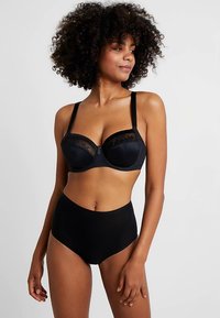 Mulher com cabelo encaracolado, vestindo um soutien preto com aro e detalhe em renda, acompanhado de cuecas pretas de cintura alta, em fundo liso.