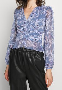 Blomstret bluse i lyseblå med V-hals, rynkedetalje, lange ballonærmer og peplumkant. Kombineret med sorte bukser i kunstlæder.