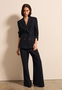 Blazer negro ajustado de doble botonadura con mangas enrolladas, combinado con pantalones de pierna ancha. Tela suave con silueta estructurada y detalles mínimos.