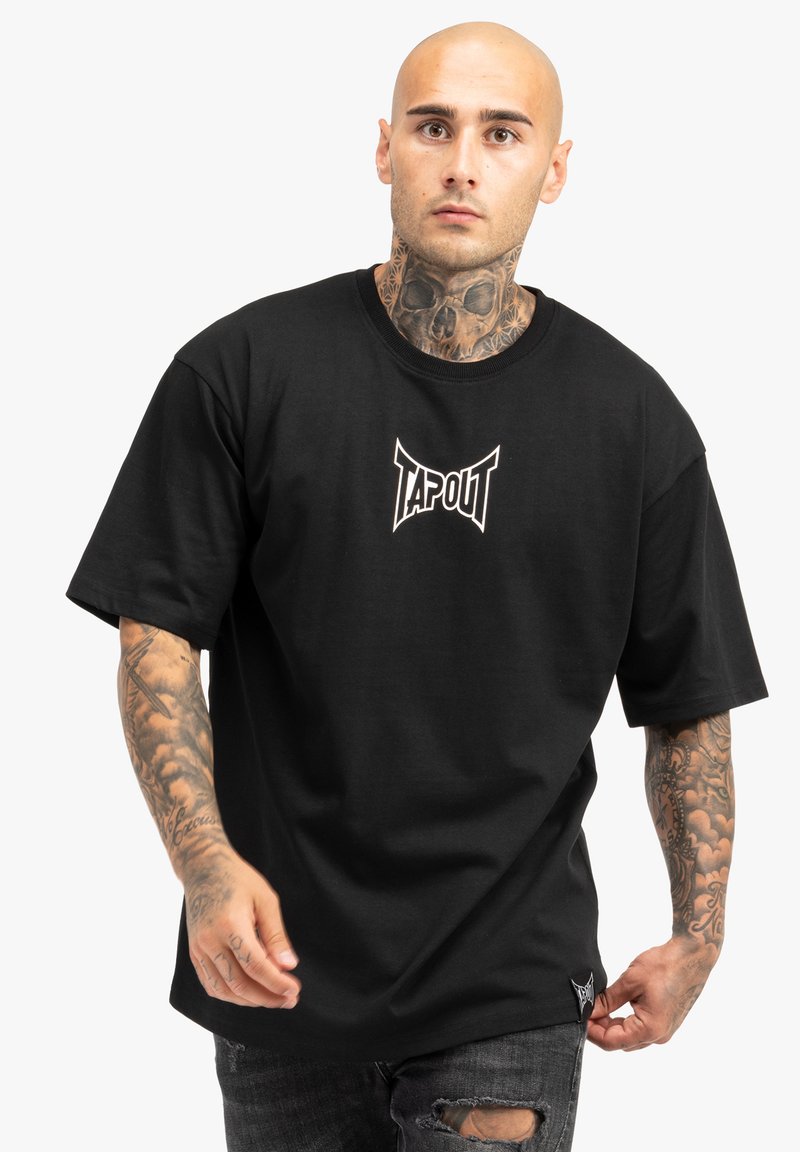 Juodas medvilnės T-marškinėlis su TAPOUT logotipu baltos spalvos, trumpomis rankovėmis, laisvu kirpimu ir matomomis siuvimo detalėmis, derinamas su susidėvėjusiais džinsais.
