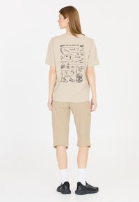 Beige t-shirt med en bakgrundsillustration av en picknickguide, kombinerad med beige knälånga shorts och svarta träningsskor.