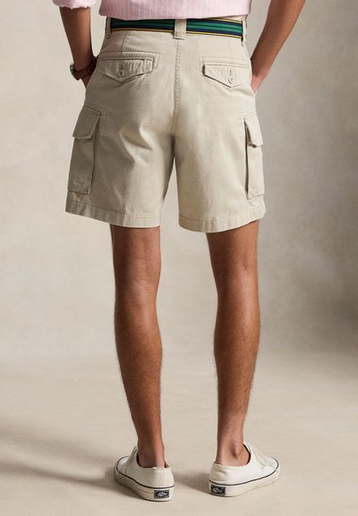 Muž nosiaci béžové cargo šortky s gombíkovými vreckami, pruhovaný opasok a biele tenisky, stojí s rukami vreckách chrbtom k nám.