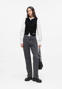 Black cropped vest over a white button-up shirt, paired with gray high-waisted jeans and black platform boots. A branded bag hangs from one hand.