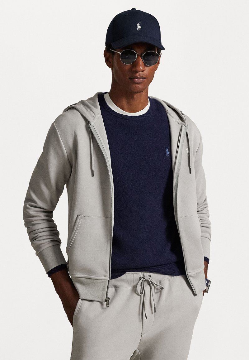 Um hoodie cinza claro com fecho éclair, apresentando cordões e bolsos, sobreposto a um suéter de gola redonda azul marinho. Os acessórios incluem um boné azul marinho e óculos de sol redondos.