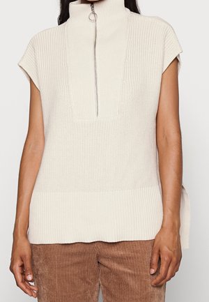 Pull en maille crème sans manches avec col montant et fermeture éclair devant, porté avec un pantalon en velours côtelé marron.