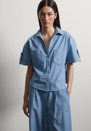 Mujer con camisa de manga corta azul clara con dos bolsillos en el pecho y pantalones sueltos a juego, de pie frente a un fondo liso.