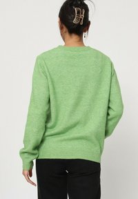 Pull vert en matériau doux, avec un col rond, des poignets et un ourlet côtelés. Le dos présente une finition texturée et une silhouette ajustée.