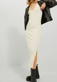 Frau in einem ärmellosen cremefarbenen, gerippten Midikleid mit seitlichem Schlitz, schwarze Lederjacke über den Schultern drapiert und schwarze klobige Stiefeletten.