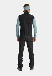 Schwarze isolierte Weste mit hohem Kragen und reflektierenden Akzenten, kombiniert mit schwarzen Hosen. Das Modell trägt ein hellblaues Langarmoberteil.