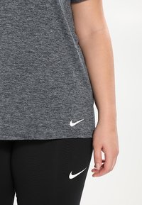 Camiseta atlética de color gris oscuro jaspeado con mangas cortas, que presenta un logo blanco de Nike en el dobladillo; combinada con leggings negros, también con logo de Nike.