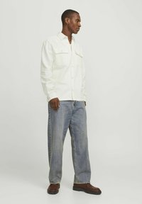 Camisa blanca de botones con dos bolsillos en el pecho, combinada con unos jeans holgados de color azul claro y zapatos marrones. La tela parece tener textura.