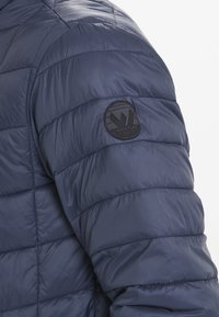 Mörkblå pufferjacka med quiltad textur, med en rund svart logotypklistermärke på den övre vänstra ärmen. Slät, lätt material.