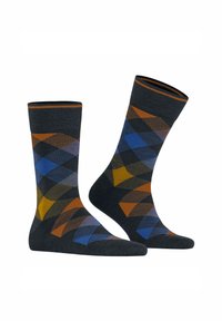 Deux chaussettes affichées debout, sur un fond sombre avec un motif argyle dans des tons de bleu, marron et jaune moutarde.